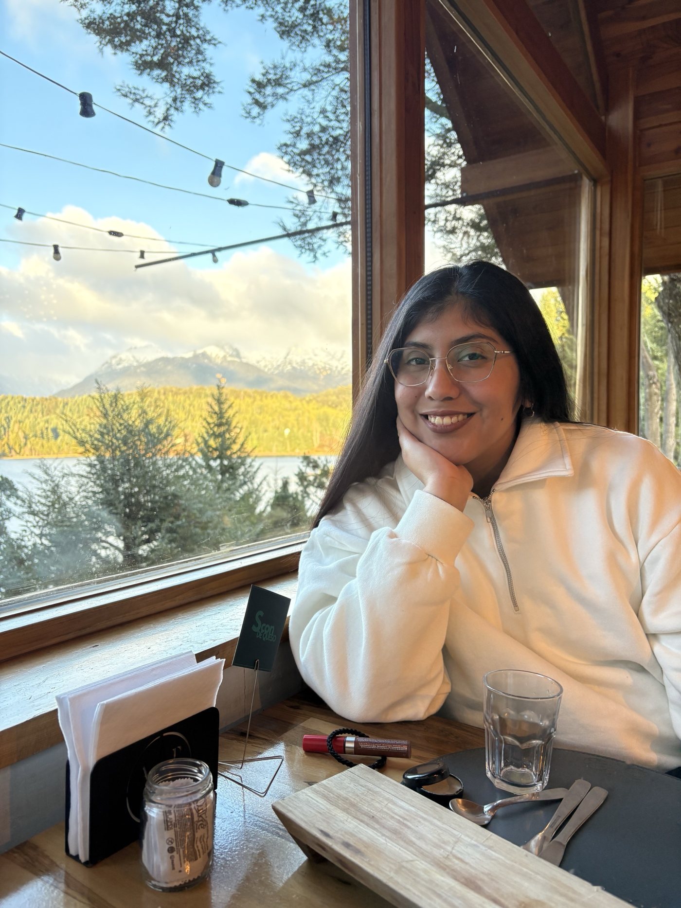 Andrea Garzon sonríe sentada junto a una ventana con vista a un lago y montañas.
